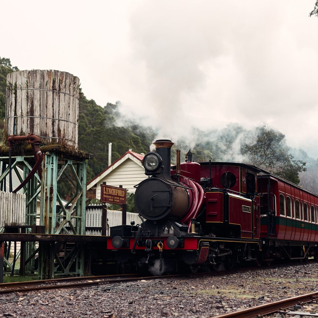 A steam locomotive waiting by the station's platform at Lynchford station, water tank in foreground, steam billowing from the locomotive's chimney.